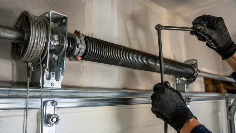 Technician's gloved hands setting a winding bar in a garage door torsion spring above the door